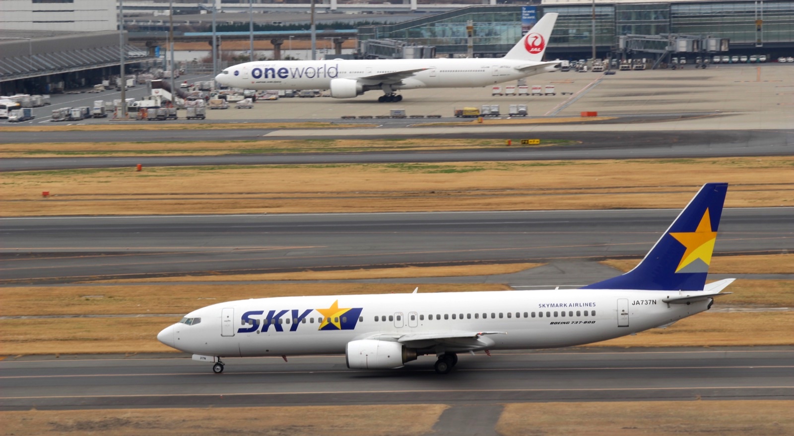 羽田空港を左へ走るスカイマークのボーイング737-800、JA737Nを横から撮った写真。奥にワンワールド塗装のJALのボーイング777が写っている
