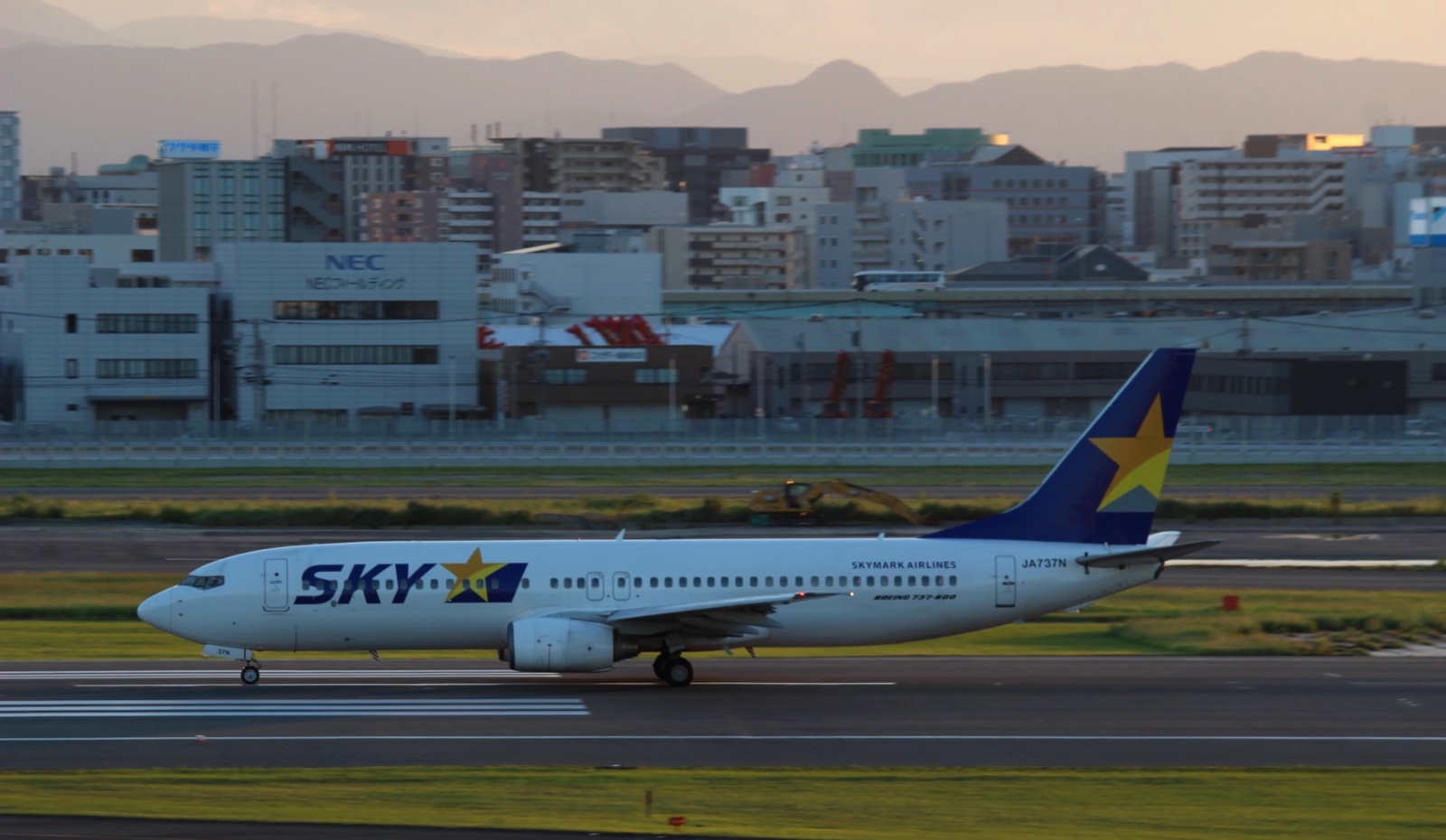 夕方の光の中、福岡空港の滑走路を走るスカイマークのJA737N。奥に福岡の街並みと山が見える