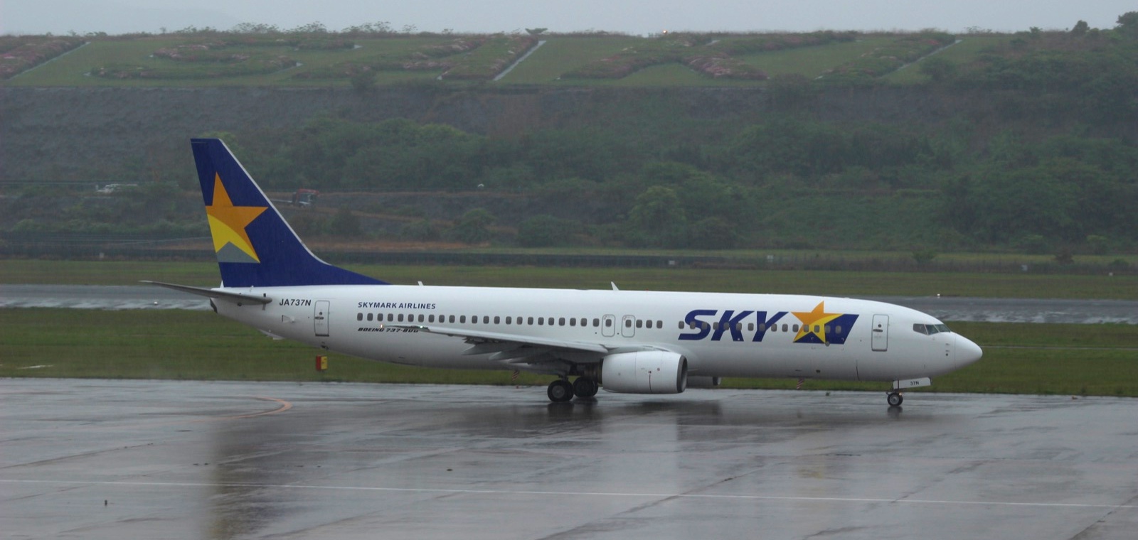 雨の長崎空港で、ぬれた地面の上を走るスカイマークのJA737N。奥に緑の丘が見える